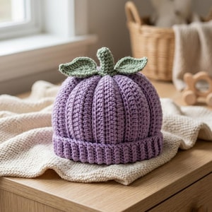 Könnte beinhalten: Eine violette Häkelmütze in Kürbisform mit gerippter Textur, einem dicken umgeschlagenen Rand und zwei hellgrünen Häkelblättern, die an einem kleinen zentralen Stiel oben auf der abgerundeten Krone befestigt sind.