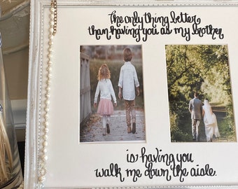 Op de afbeelding: Een witte, verweerde houten fotolijst met twee foto's en de zwarte cursieve tekst: The only thing better than having you as my brother is having you walk me down the aisle. Een kralenkwastje hangt aan de lijst.