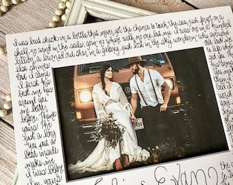 May include: A white rectangular picture frame featuring handwritten black cursive text surrounding a central photograph of a couple standing in front of a vintage orange van. The text includes the names Tali & Evan and poetic verses.