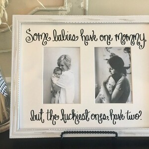 May include: White double photo frame with the text "Some babies have one mommy, but the luckiest ones have two?" in black script. The frame holds two black and white photos of a mother holding a baby. The frame is on a black stand.