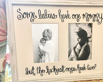 Op de afbeelding: Een witte fotolijst met twee zwart-witfoto's van moeders die baby's vasthouden. De tekst luidt: "Sommige baby's hebben één mama, maar de gelukkigste hebben er twee?" De lijst heeft een verweerde afwerking.