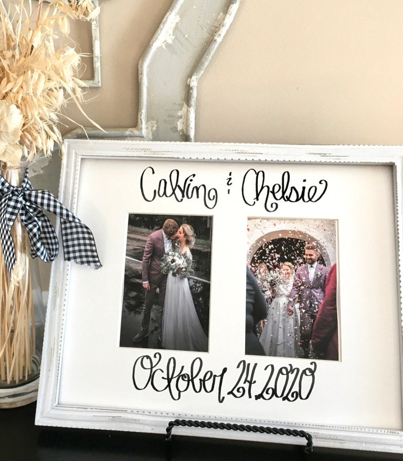 Op de afbeelding: Een wit fotokader met twee trouwfoto's. De namen "Calvin & Chelsie" en de datum "24 oktober 2020" zijn in zwart schrift geschreven. Het frame wordt ondersteund door een zwarte metalen standaard.