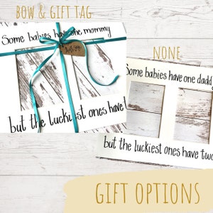 May include: White framed artwork with the text "Some babies have one mommy but the luckiest ones have..." and "Some babies have one daddy but the luckiest ones have two". A teal ribbon and gift tag are attached.