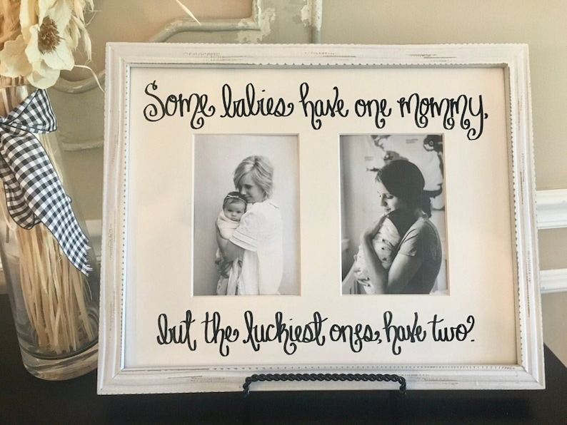 May include: White double photo frame with the text "Some babies have one mommy, but the luckiest ones have two?" in black script. The frame holds two black and white photos of a mother holding a baby. The frame is on a black stand.