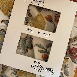 Op de afbeelding: Een witte fotolijst met twee openingen, met de tekst "Like father..." en "Like son." De lijst bevat de jaren "1976" en "2021" met een hartsymbool. De achtergrond toont een bloemenstof.