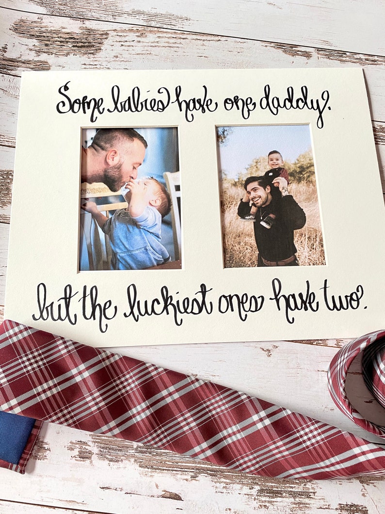 May include: A cream-colored photo frame with two openings, featuring the text "Some babies have one daddy, but the luckiest ones have two." A red and gray plaid tie is in the foreground.