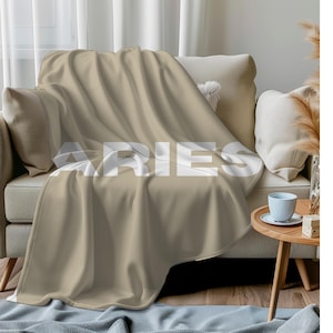 May include: A solid beige throw blanket draped over a sofa, featuring the word ARIES printed in large, bold, white capital letters across the center of the fabric. The blanket has a smooth, uniform texture and covers the seat and back cushions.
