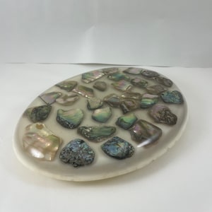 Puede incluir: Posavasos ovalado blanco con un mosaico de piezas de concha iridiscente verde, azul y blanca incrustadas en la superficie.