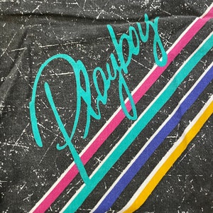Peut inclure: Un t-shirt noir avec le mot "Playboy" &eacute;crit en turquoise sur le devant. Le t-shirt a des rayures color&eacute;es en rose, turquoise, bleu et jaune.