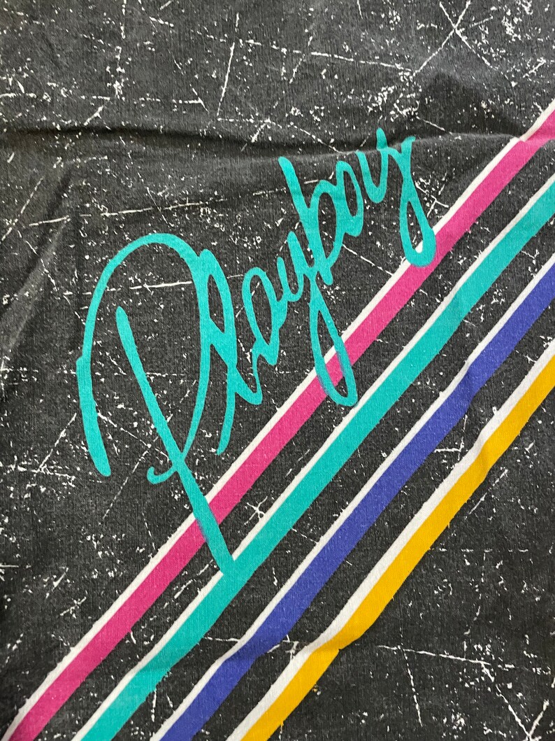 Peut inclure: Un t-shirt noir avec le mot "Playboy" &eacute;crit en turquoise sur le devant. Le t-shirt a des rayures color&eacute;es en rose, turquoise, bleu et jaune.