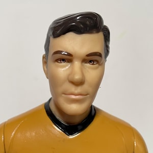 May include: A vintage action figure of a man with brown hair and brown eyes, wearing a yellow uniform with a black collar and a black and white emblem. The figure has a serious expression.