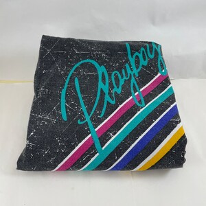 Peut inclure: Un t-shirt noir &agrave; l'aspect vieilli avec un graphisme turquoise du mot "Playboy" avec des rayures color&eacute;es derri&egrave;re.