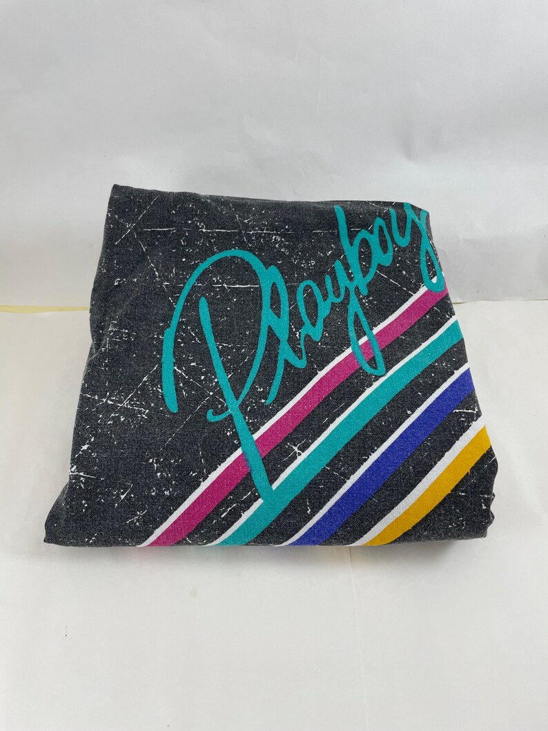 Peut inclure: Un t-shirt noir &agrave; l'aspect vieilli avec un graphisme turquoise du mot "Playboy" avec des rayures color&eacute;es derri&egrave;re.