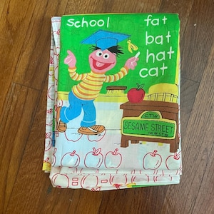 Puede incluir: Una sábana de cama de Barrio Sésamo doblada con una ilustración de dibujos animados de un personaje con un gorro de graduación. La sábana tiene las palabras "SCHOOL" y "fat bat hat cat" impresas. La sábana es blanca con diseños de manzanas rojas.