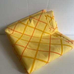 May include: Folded yellow fabric with a grid pattern of orange and yellow lines. The fabric is folded over itself, creating a layered effect. The background is a plain white surface.