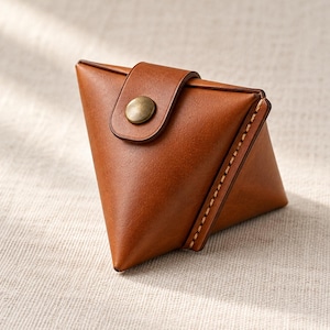 May include: A triangular coin pouch crafted from smooth brown leather, featuring visible light-colored stitching along the edges and a single metallic snap button closure centered on the front flap of the small accessory.