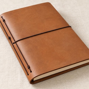 May include: A tan notebook with a smooth cover, secured by a dark brown elastic cord that wraps around the center and threads through two holes on the spine. The notebook contains off-white pages visible along the side edge.