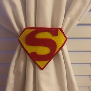 Peut inclure: Un attache-rideau en feutre en forme de logo Superman. Il présente un diamant jaune avec un bord rouge et un "S" rouge au centre. L'attache-rideau maintient des rideaux blancs.