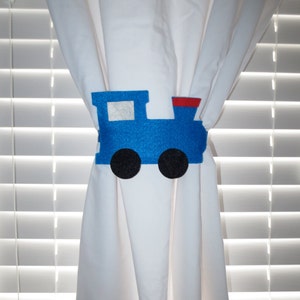 May include: Blue felt train curtain tieback with black wheels and a red chimney. The tieback is attached to a white curtain.
