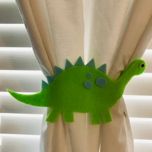 May include: Green felt dinosaur curtain tieback with blue accents. The dinosaur has a long tail and a spiky back.