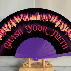 May include: A purple folding hand fan with a black background and the words "GASH YOUR TEETH" in red. The background is decorated with a pattern of teeth.
