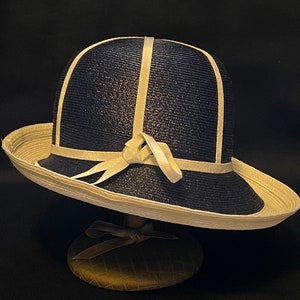 May include: A navy blue and cream-colored hat with a rounded crown and a wide brim. The hat features cream-colored trim and a decorative bow. The hat is displayed on a hat stand against a dark background.