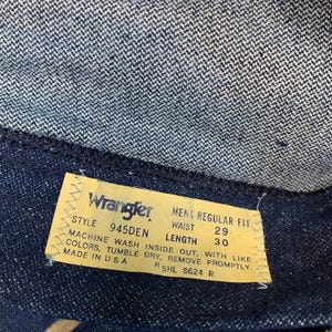 May include: Close-up of a Wrangler jeans label. The yellow label has black text detailing the style, waist size 74 cm, and length 76 cm. The jeans are dark blue denim. The label also states "Made in USA".