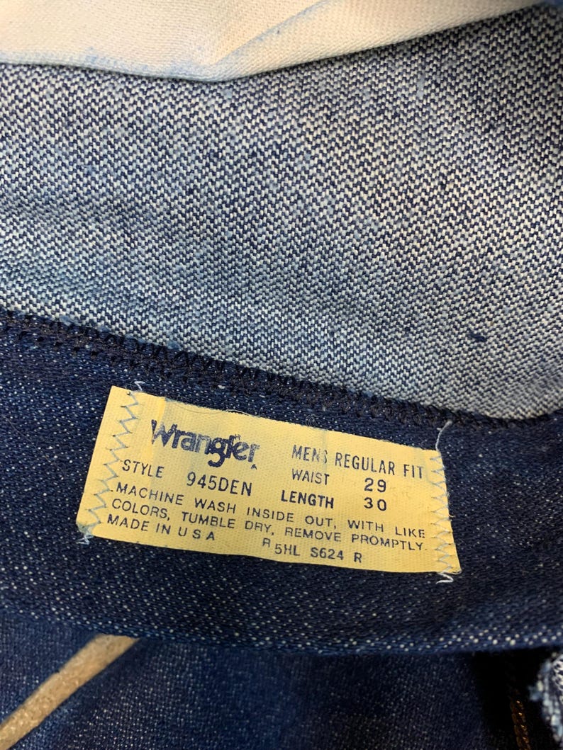 May include: Close-up of a Wrangler jeans label. The yellow label has black text detailing the style, waist size 74 cm, and length 76 cm. The jeans are dark blue denim. The label also states "Made in USA".