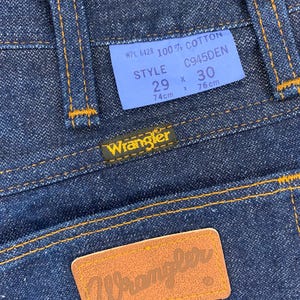 May include: Close-up of dark blue denim jeans with orange stitching. A blue tag displays "WPL 6428 100% COTTON" and size details: 29 x 30, with 74cm x 76cm measurements. The Wrangler logo is visible on a leather patch.
