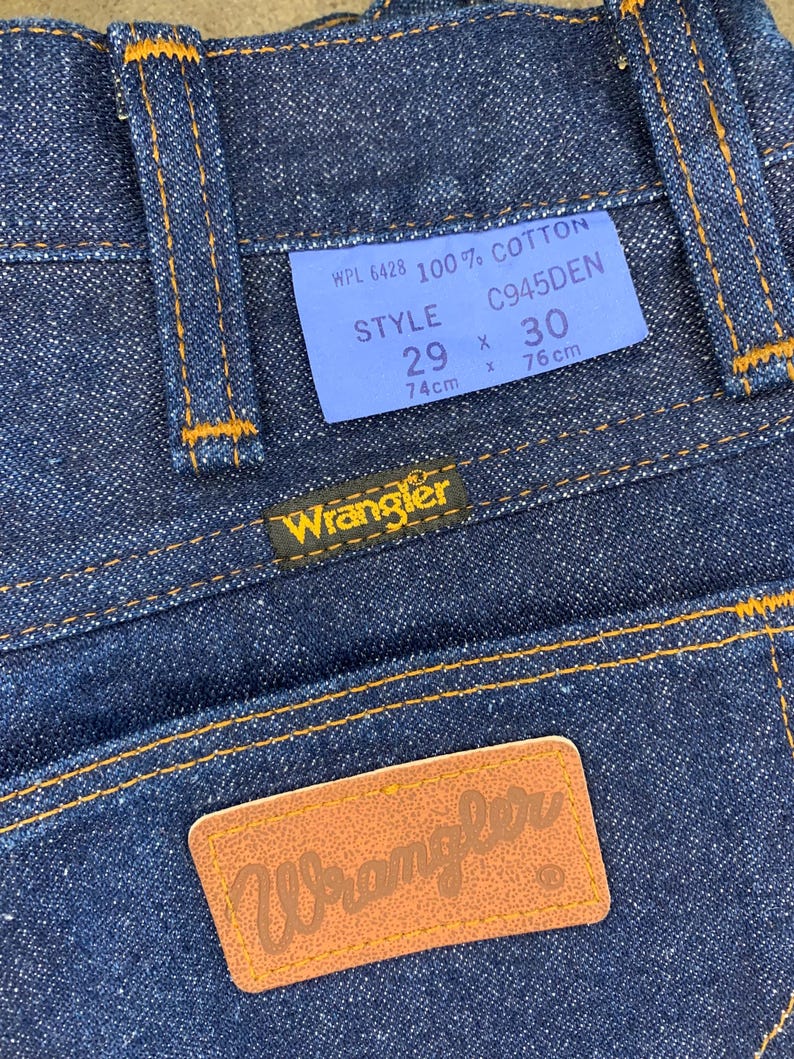 May include: Close-up of dark blue denim jeans with orange stitching. A blue tag displays "WPL 6428 100% COTTON" and size details: 29 x 30, with 74cm x 76cm measurements. The Wrangler logo is visible on a leather patch.