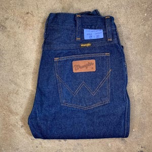 May include: Dark blue Wrangler jeans with a classic five-pocket design. The jeans feature a brown leather patch with the Wrangler logo and a blue tag with the size 29 x 30. The jeans are made of 100% cotton.