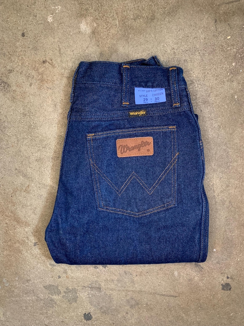 May include: Dark blue Wrangler jeans with a classic five-pocket design. The jeans feature a brown leather patch with the Wrangler logo and a blue tag with the size 29 x 30. The jeans are made of 100% cotton.