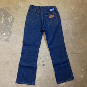 May include: Dark blue denim jeans with a classic five-pocket design. Features the Wrangler logo on the back pocket and a blue tag. The jeans have a straight leg cut.