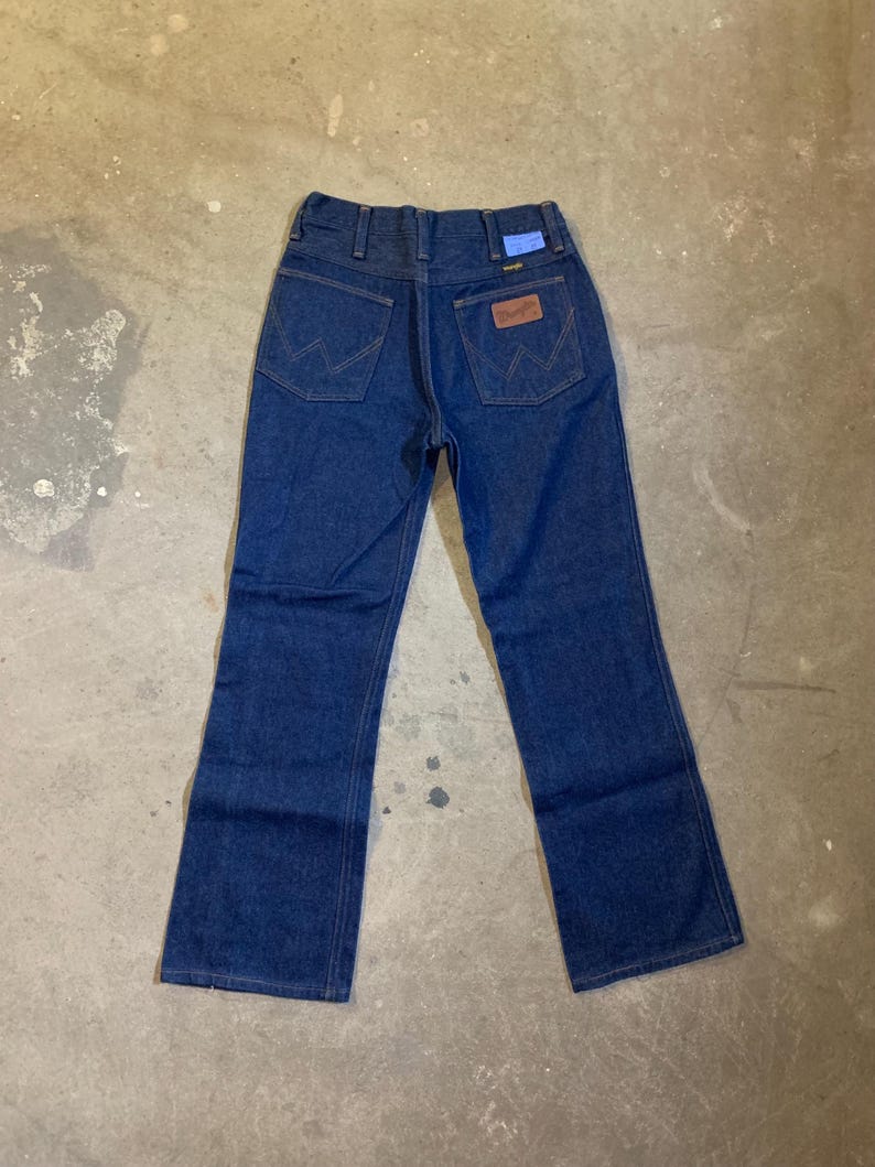May include: Dark blue denim jeans with a classic five-pocket design. Features the Wrangler logo on the back pocket and a blue tag. The jeans have a straight leg cut.