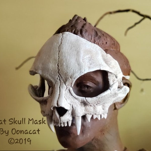 May include: A white cat skull mask with a detailed bone structure and sharp teeth. The mask is worn on a mannequin head.