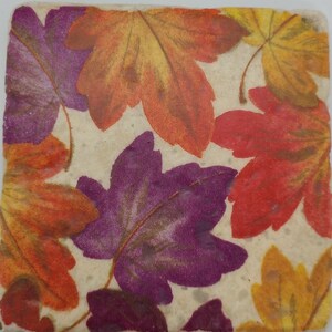 May include: A square coaster with a white background and a pattern of colorful fall leaves. The leaves are in shades of orange, red, purple, and yellow.