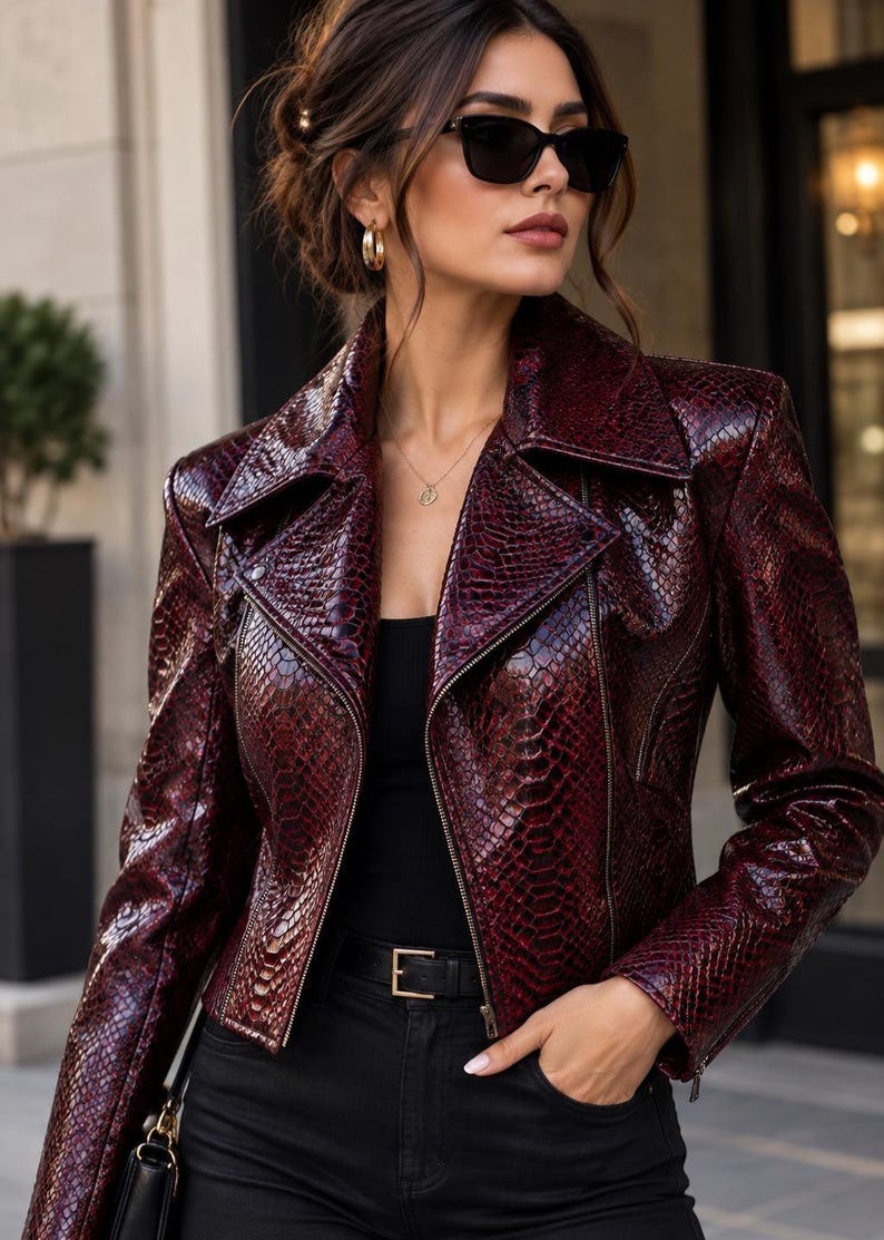 Puede incluir: Una chaqueta motera de color burdeos oscuro con un patr&oacute;n de piel de serpiente texturizado, cuello de solapa ancho y cremalleras plateadas. La chaqueta se lleva sobre una camiseta negra lisa y pantalones negros con un cintur&oacute;n de hebilla dorada.