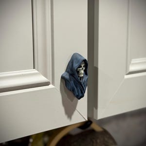 May include: A cabinet knob shaped like a hooded figure with a visible skull face. The figure is colored in shades of dark blue and grey, featuring a detailed skeletal structure with deep eye sockets and a bared jaw, mounted on a white cabinet door.