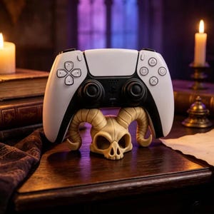 May include: A white and black wireless video game controller rests on a beige, bone-colored stand shaped like a horned skull. The controller features a central touchpad, two analog sticks, a directional pad, and four action buttons with symbols.