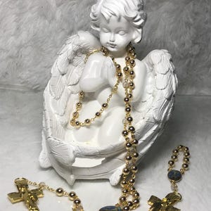 May include: A white cherub statue with gold rosary beads draped around it. The rosary features gold beads and a gold cross pendant. A smaller rosary with a blue stone is also present.