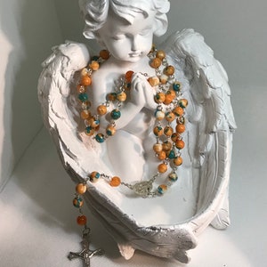 May include: A white cherub figurine with wings holding a multi-strand rosary with orange and turquoise beads. The rosary has a silver cross.
