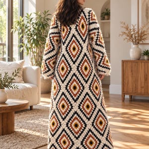 May include: A floor-length crochet duster cardigan featuring a repeating diamond pattern in cream, black, rust orange, and mustard yellow. The garment has long, loose sleeves and a scalloped hem, constructed from thick, textured yarn.