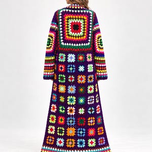 Può includere: Un cappotto lungo all'uncinetto caratterizzato da un vivace patchwork di granny square multicolori su uno sfondo viola intenso. Il capo presenta maniche lunghe e un motivo ripetuto di piccoli quadrati colorati su tutto il design.