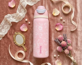 Könnte beinhalten: Eine hellrosa Owala-Wasserflasche mit weißem Blumenmuster, einem Druckknopfdeckel mit Verriegelungsmechanismus und dem vertikal in Weiß aufgedruckten Markennamen. Die Flasche ist umgeben von Spitzenband, einem kleinen goldenen Handspiegel und Trockenblumen.