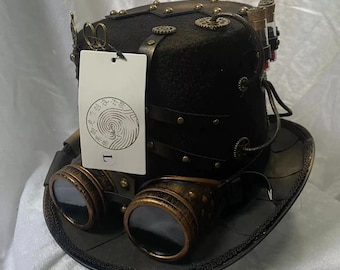 Può includere: Un cilindro nero con occhiali color ottone, ingranaggi metallici decorativi e due piccole fiale di vetro sul lato. Un'etichetta rettangolare bianca con un logo circolare e la lettera L è attaccata alla parte anteriore del cappello.