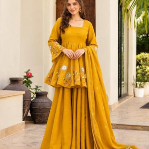 May include: A mustard yellow peplum-style kurta with long sleeves and wide-leg sharara pants. The garment features floral embroidery and circular mirror-work accents on the bodice and sleeves, paired with a matching mustard yellow dupatta.
