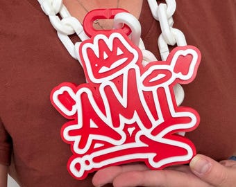 May include: A large red and white pendant featuring the word AMI in a graffiti-style font with a crown icon above it. The pendant is attached to a thick, chunky white plastic chain necklace that rests around the neck area.