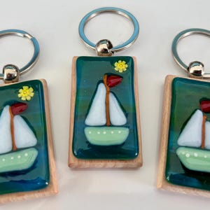 May include: Three keyrings with a sailboat design. Each keyring features a rectangular wooden frame with a teal and blue background. A white sailboat with a red sail and a yellow flower are on each keyring.