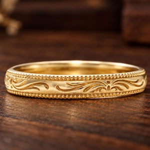 Könnte beinhalten: Ein goldener Ring mit polierter Oberfläche und eingravierten Rankenmustern auf dem Mittelband, der an der Ober- und Unterseite von feinen, perlenartigen Kanten an diesem kreisförmigen Schmuckstück aus Metall gesäumt ist.