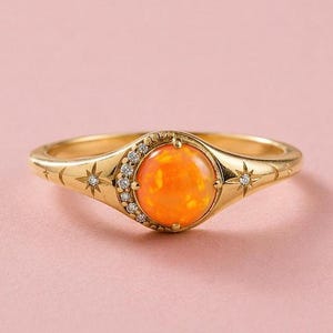 Könnte beinhalten: Ein goldener Ring mit einem runden, leuchtend orangefarbenen Opal-Cabochon in der Mitte. Die Fassung ist mit einer halbmondförmigen Reihe kleiner weißer Diamanten und eingravierten Sternmotiven auf der Ringschiene verziert.